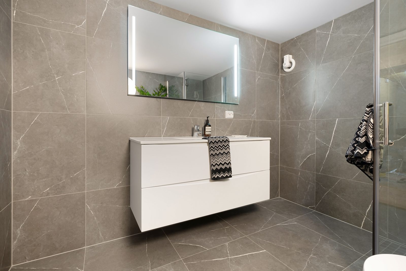 Foto de archivo. Baño con porcelánico gris, mueble suspendido blanco y ducha de cristal.