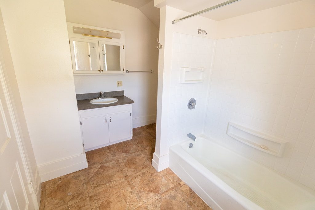 Foto de archivo. Baño antes de la reforma: mueble blanco, suelo de baldosa marrón y bañera con azulejo blanco. Es la misma estancia que la imagen de al lado.