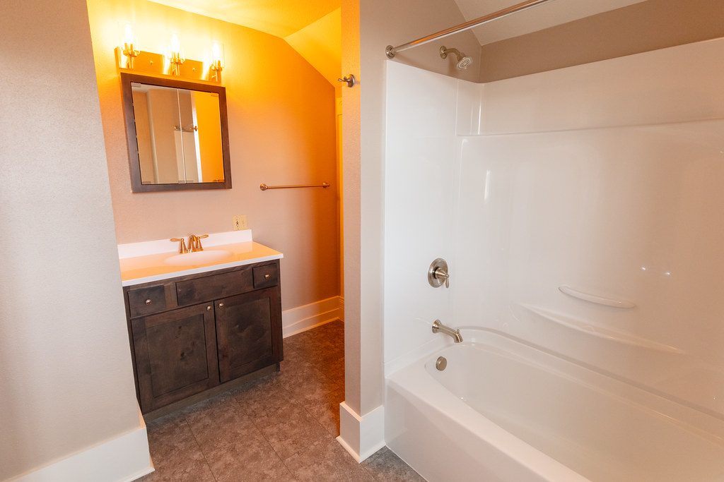Foto de archivo. El mismo baño después: mueble de madera oscura, encimera clara y bañera con frontal blanco.