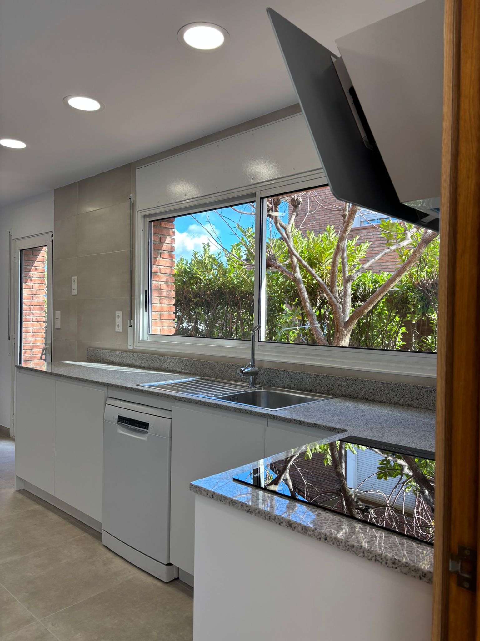 Cocina con muebles blancos, encimera de granito gris y fregadero bajo ventana.